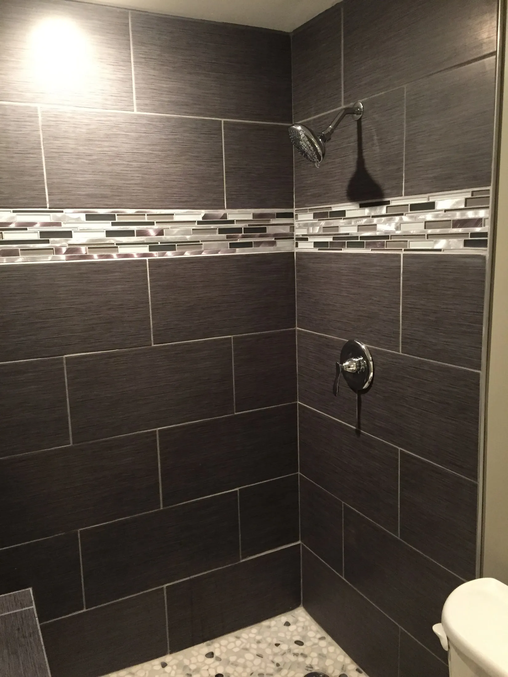 Charcoal large-format tile shower with a glass mosaic accent band and pebble floor.