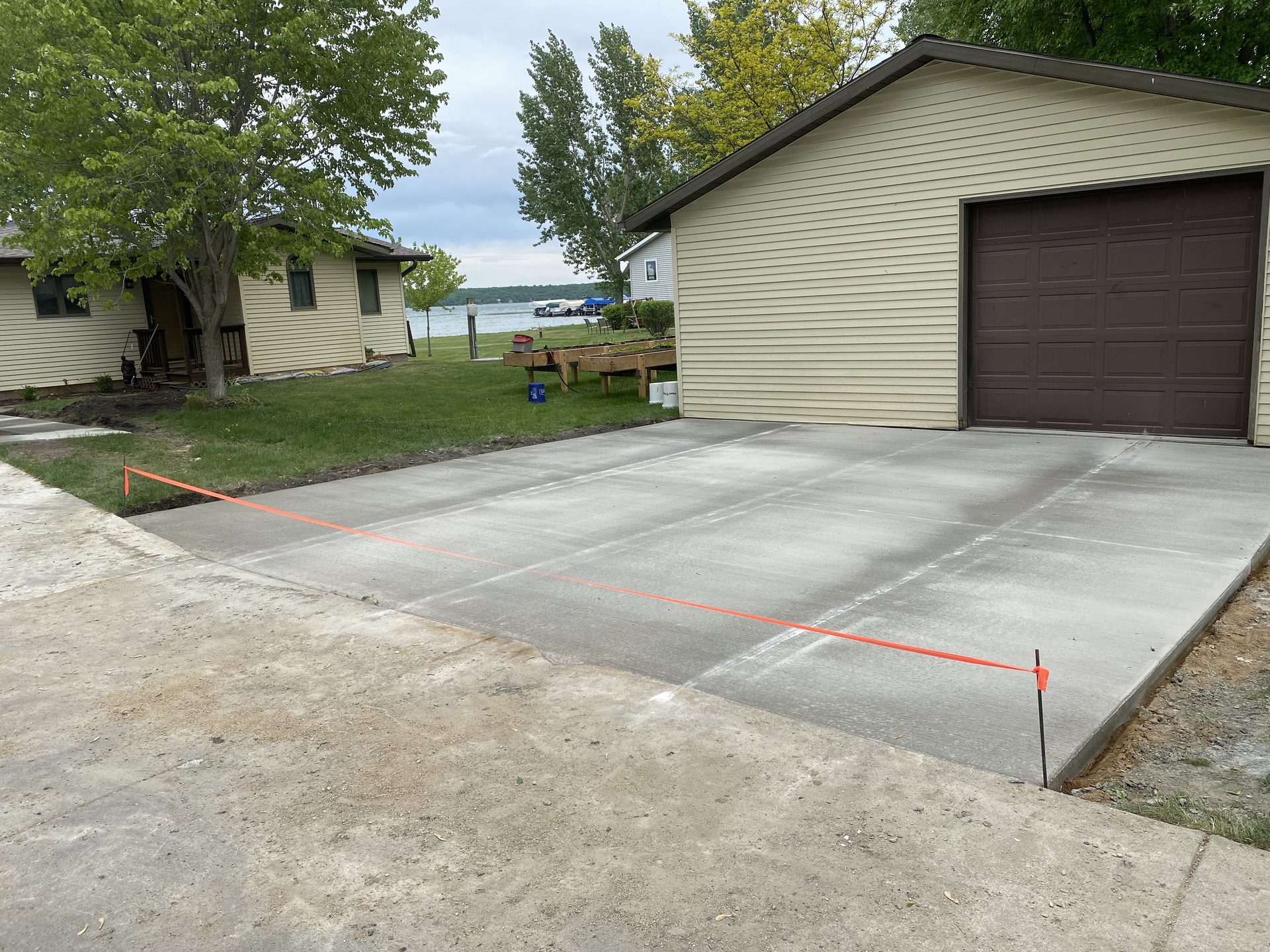 Newly poured concrete driveway adjacent to garage with orange survey line visible, backed by residential property with lake view and mature trees.