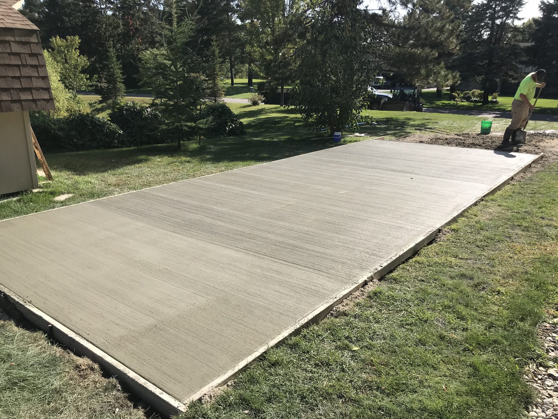 Newly poured concrete pad in residential backyard with green lawn and trees in background.