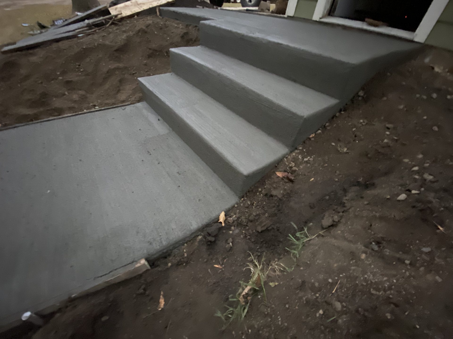 Three newly finished concrete steps leading to an entrance, with freshly poured gray concrete visible against surrounding soil.