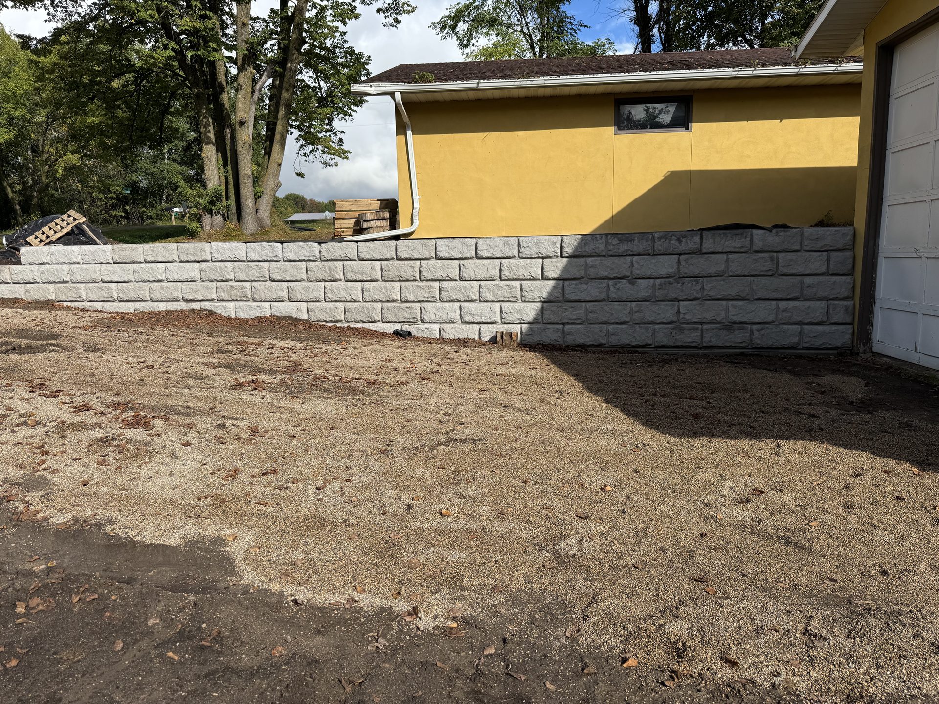 Newly constructed concrete retaining wall with yellow house and garage in background, gravel driveway area under development.