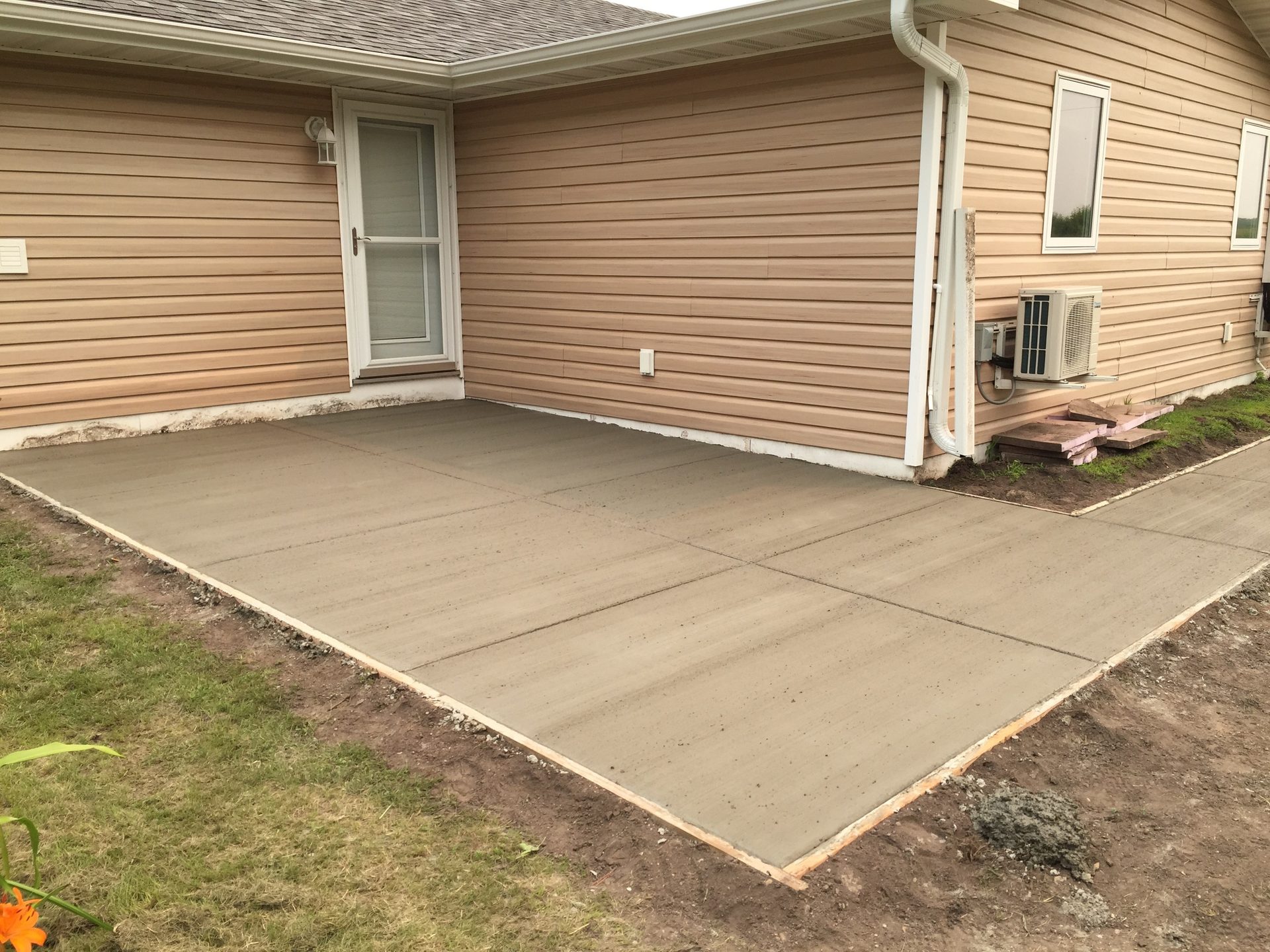 Newly finished concrete driveway adjacent to a tan vinyl-sided house with white trim and downspouts.