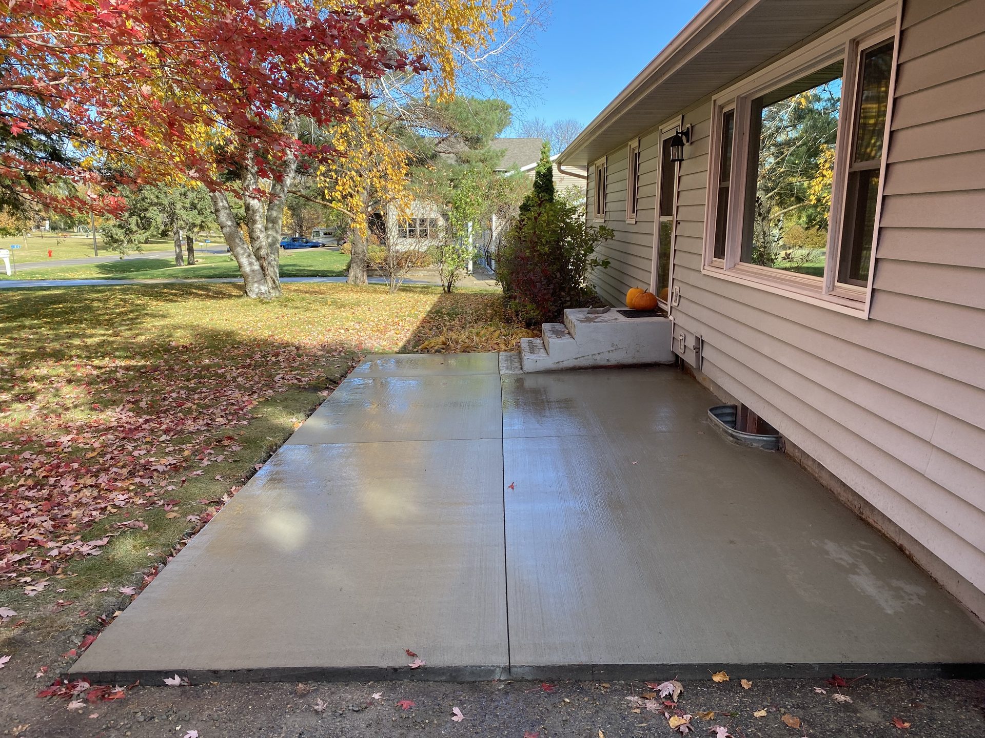 Newly completed concrete driveway extending from residential home in autumn, with fall foliage visible in background.