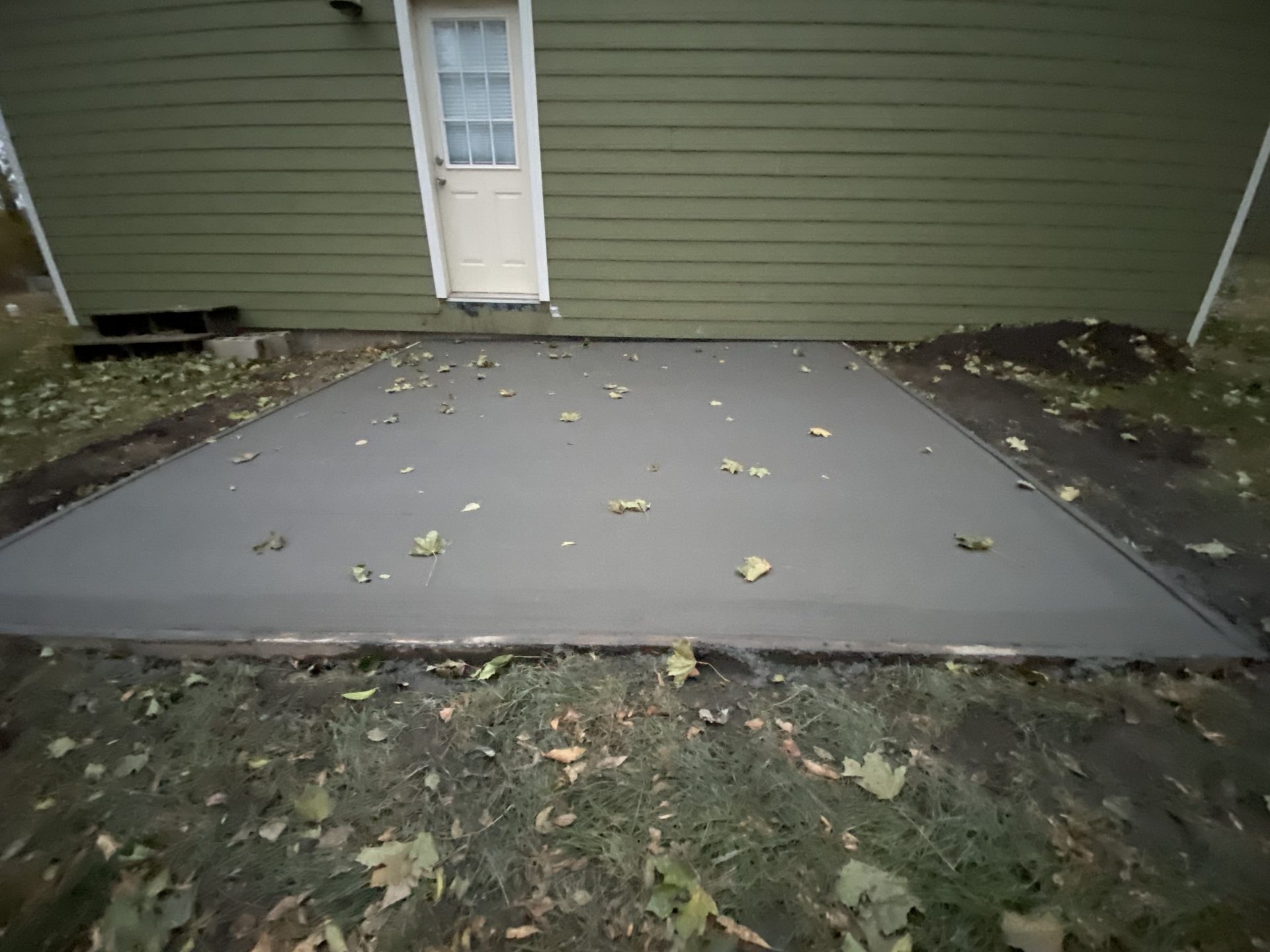 Newly installed dark gray concrete patio with fallen leaves, bordered by grass and green house with white door.