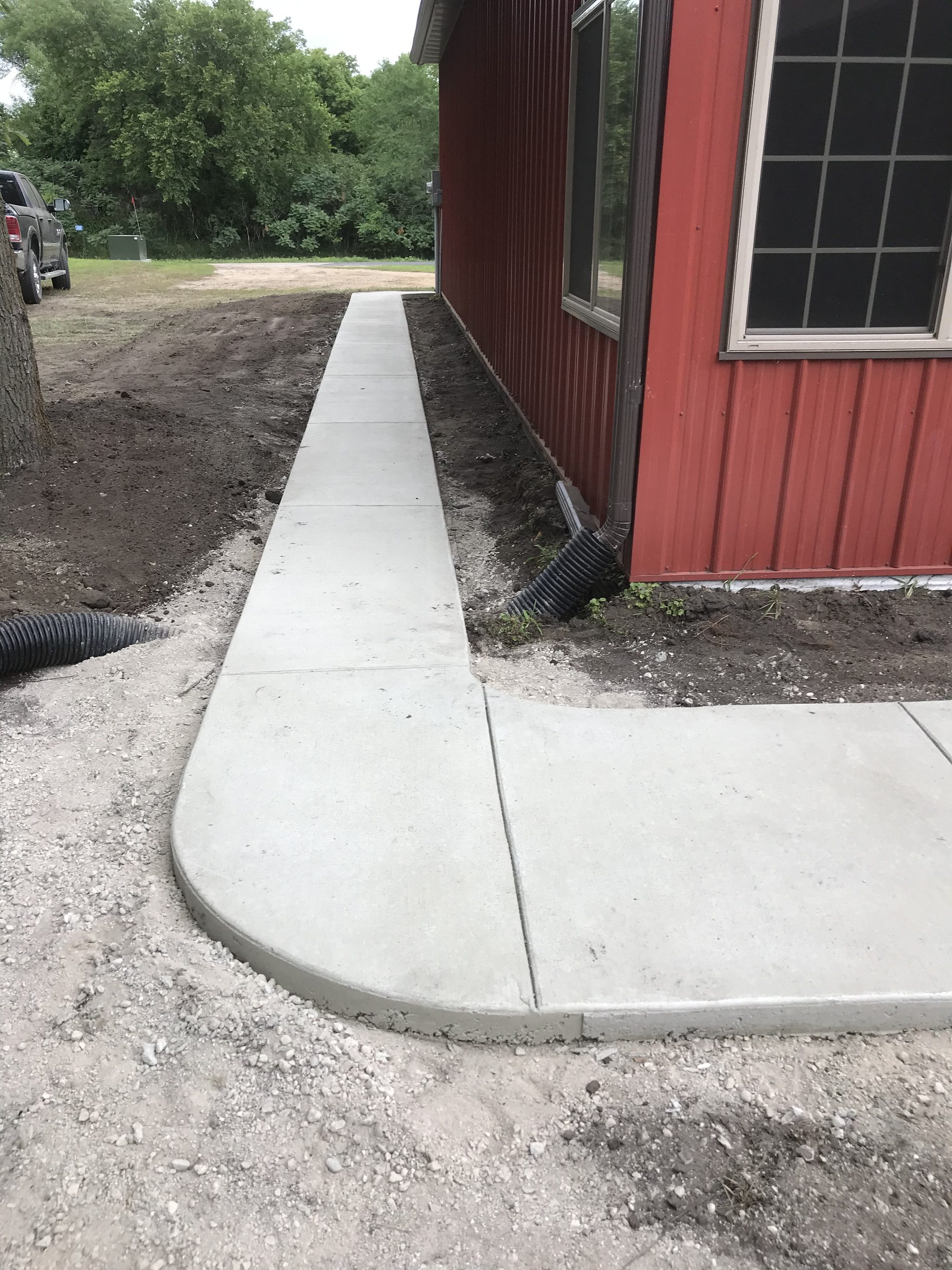 Newly installed concrete sidewalk and pad along red metal building, with curved transition detail visible.
