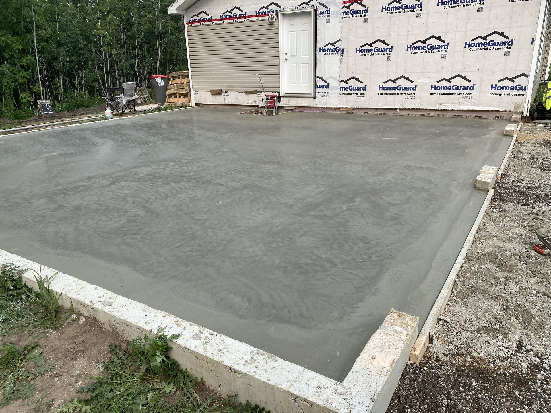 Freshly finished concrete foundation or pad with white perimeter edging, surrounded by construction site materials and HomeGuard weather protection on adjacent building structure.