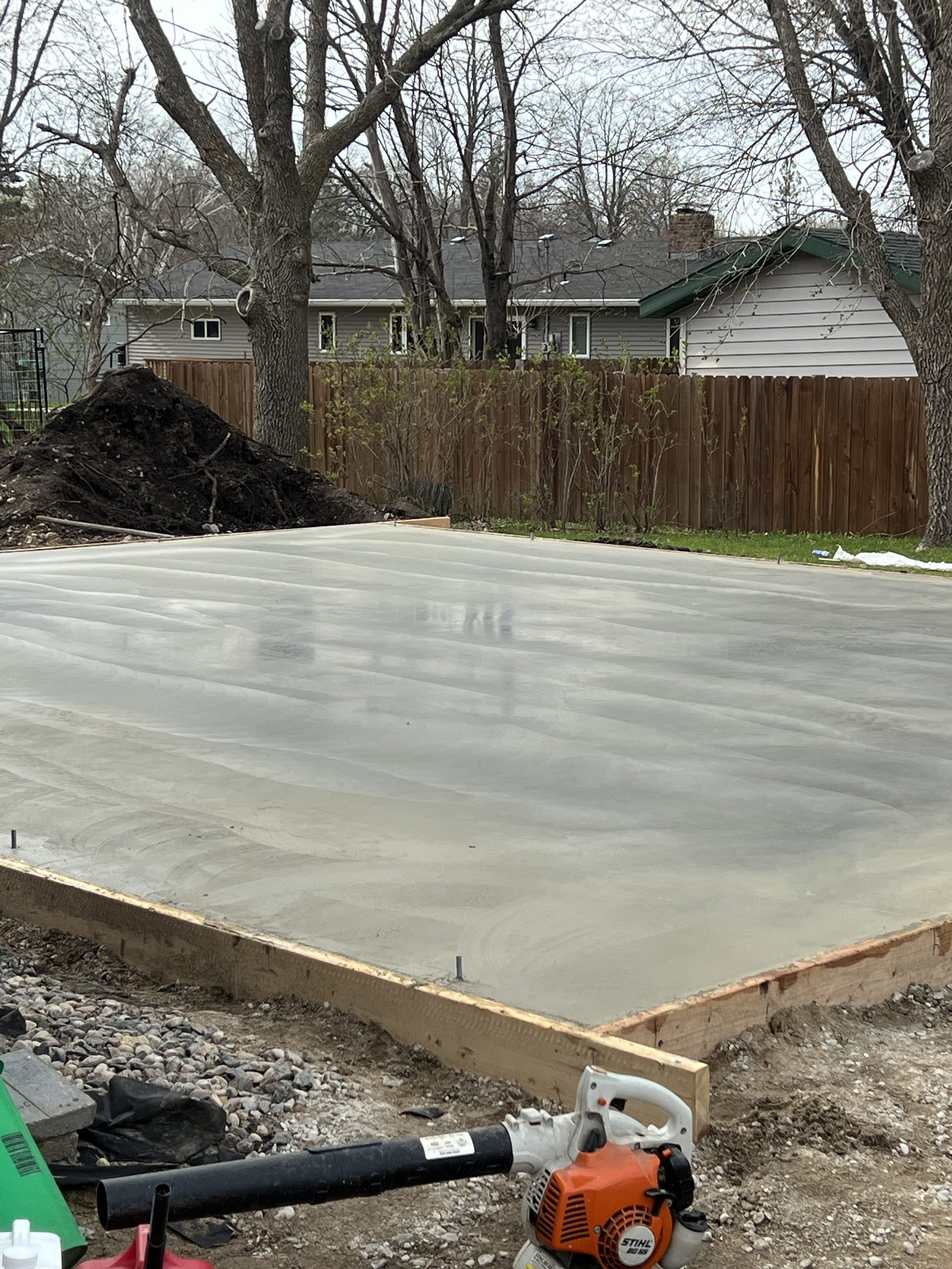 Freshly poured concrete pad with wooden frame and gravel base, surrounded by bare trees and wooden fence, showing concrete driveway foundation work.