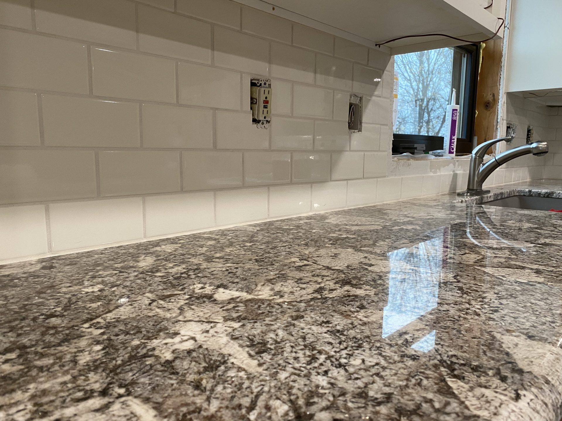 Completed kitchen backsplash with white subway tile and granite countertop with chrome faucet, side-lit with natural window light.