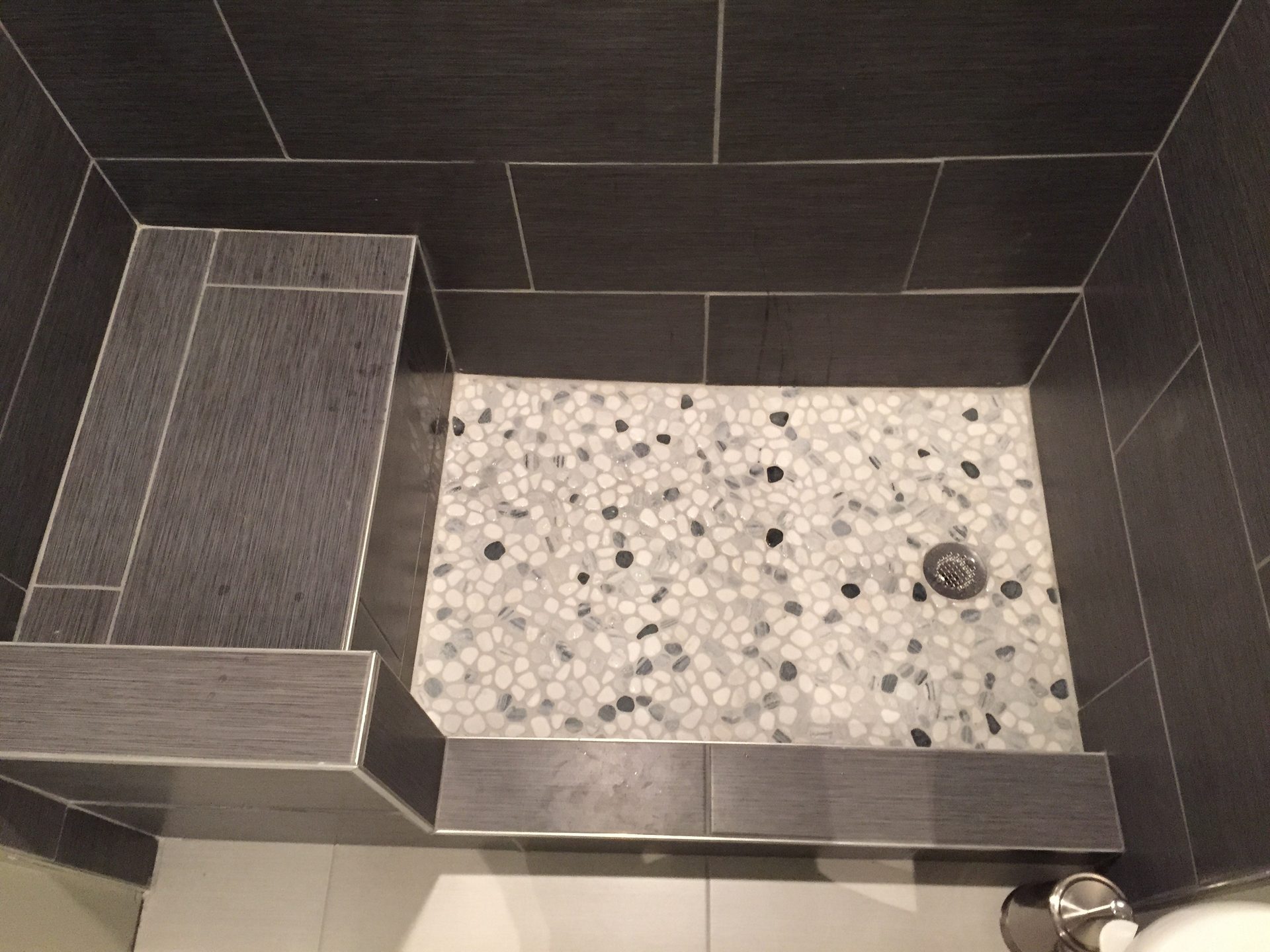 Overhead view of a completed tile shower floor featuring white hexagonal tile with black and gray accent stones and a chrome drain fixture.