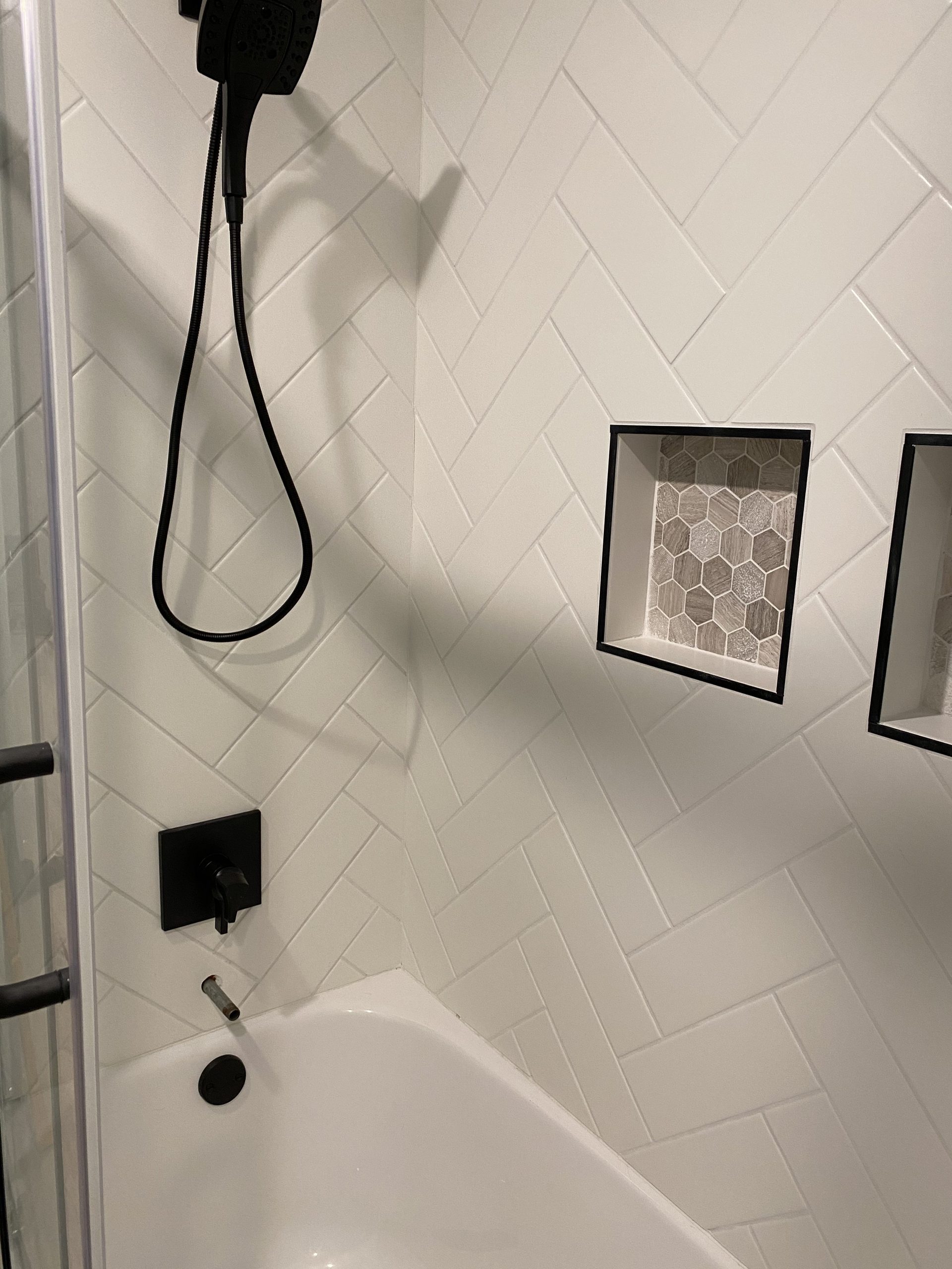 Completed tile shower featuring white subway tile in herringbone pattern with black matte fixtures and hexagonal tile niche inserts.