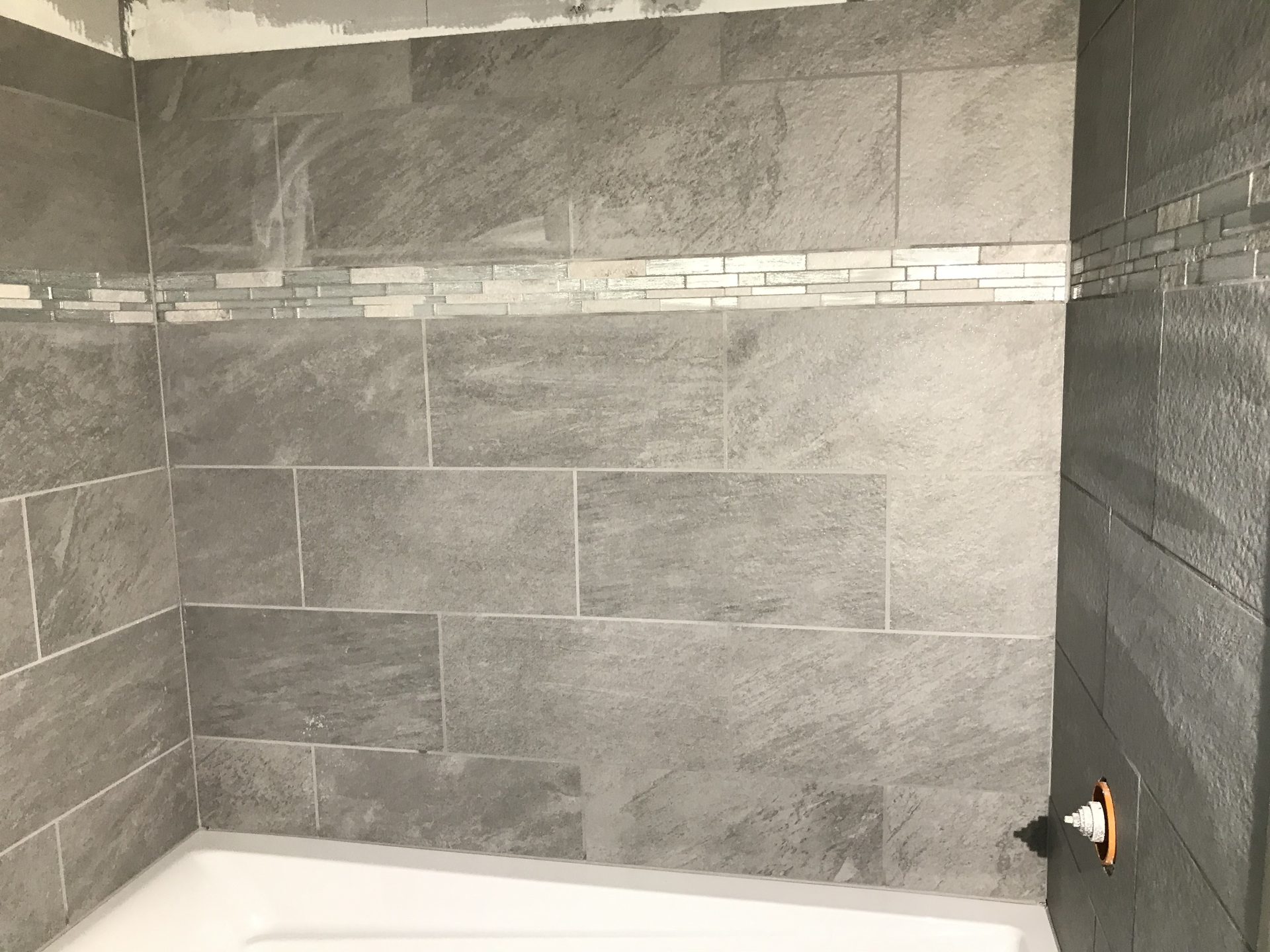 Completed tile shower surround featuring large-format gray stone-look ceramic tiles with white subway tile accent strip, installed in running bond pattern.
