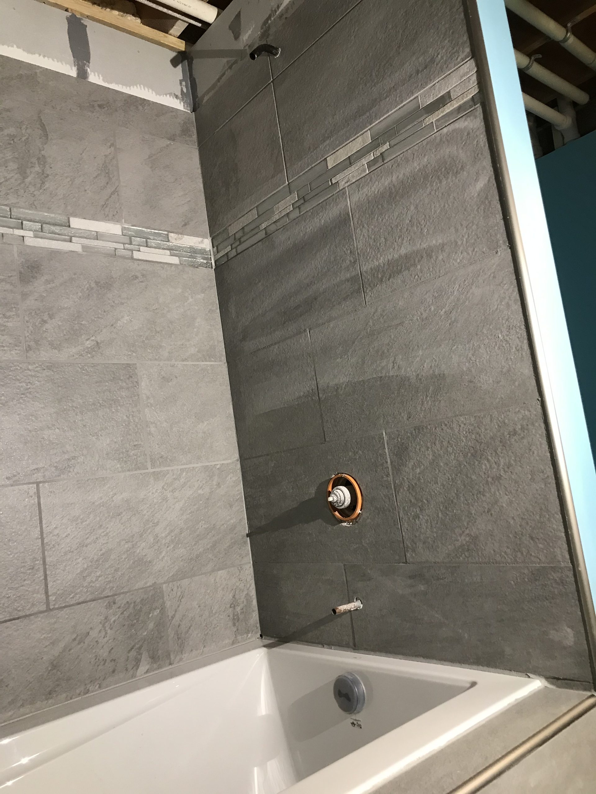 Completed tile shower installation with gray ceramic tile, decorative accent stripe, oil-rubbed bronze showerhead, and white bathtub base.