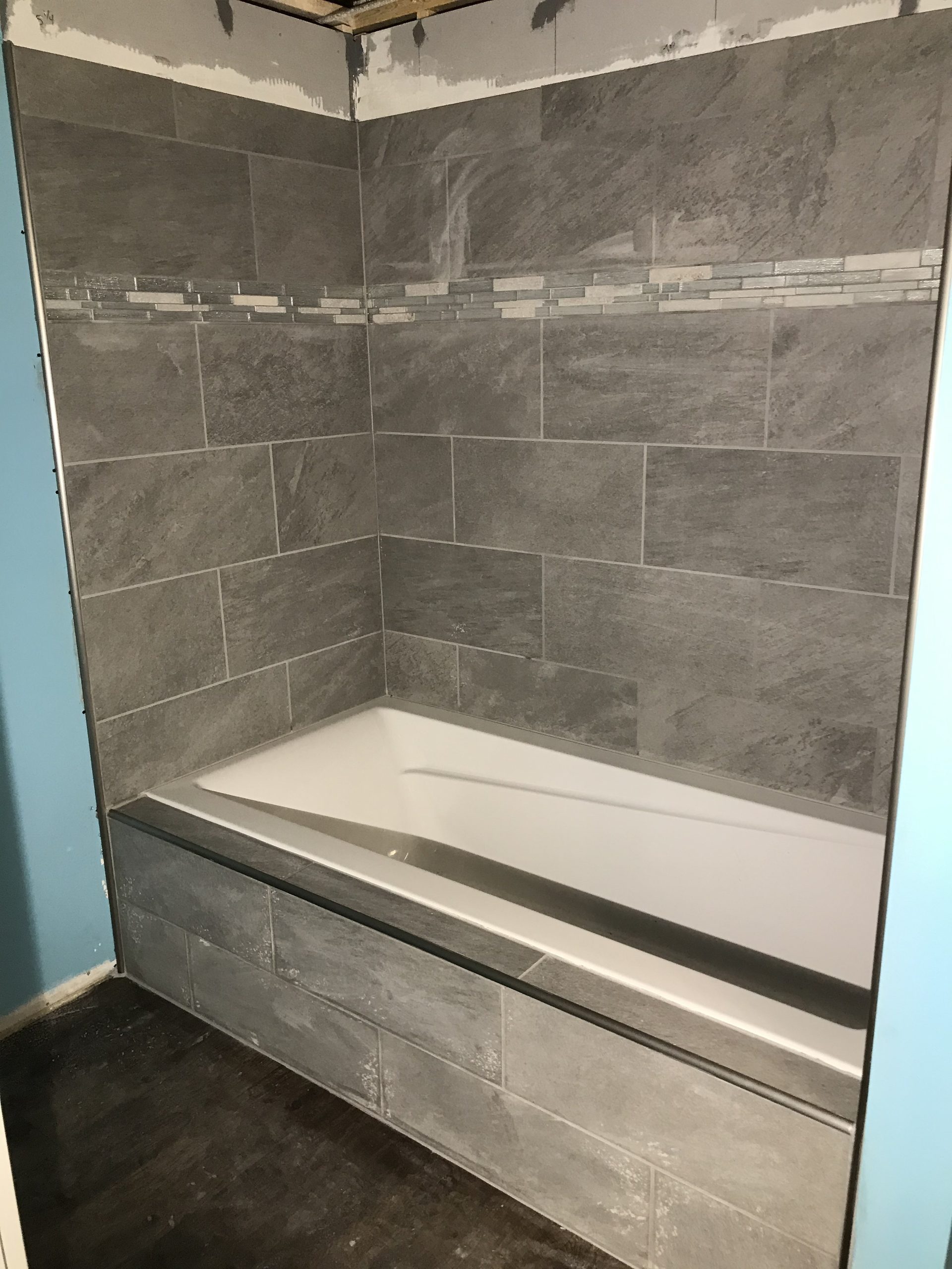 Finished tile shower surround with gray stone-look tile, linear accent strip, white acrylic tub, and frameless glass enclosure.