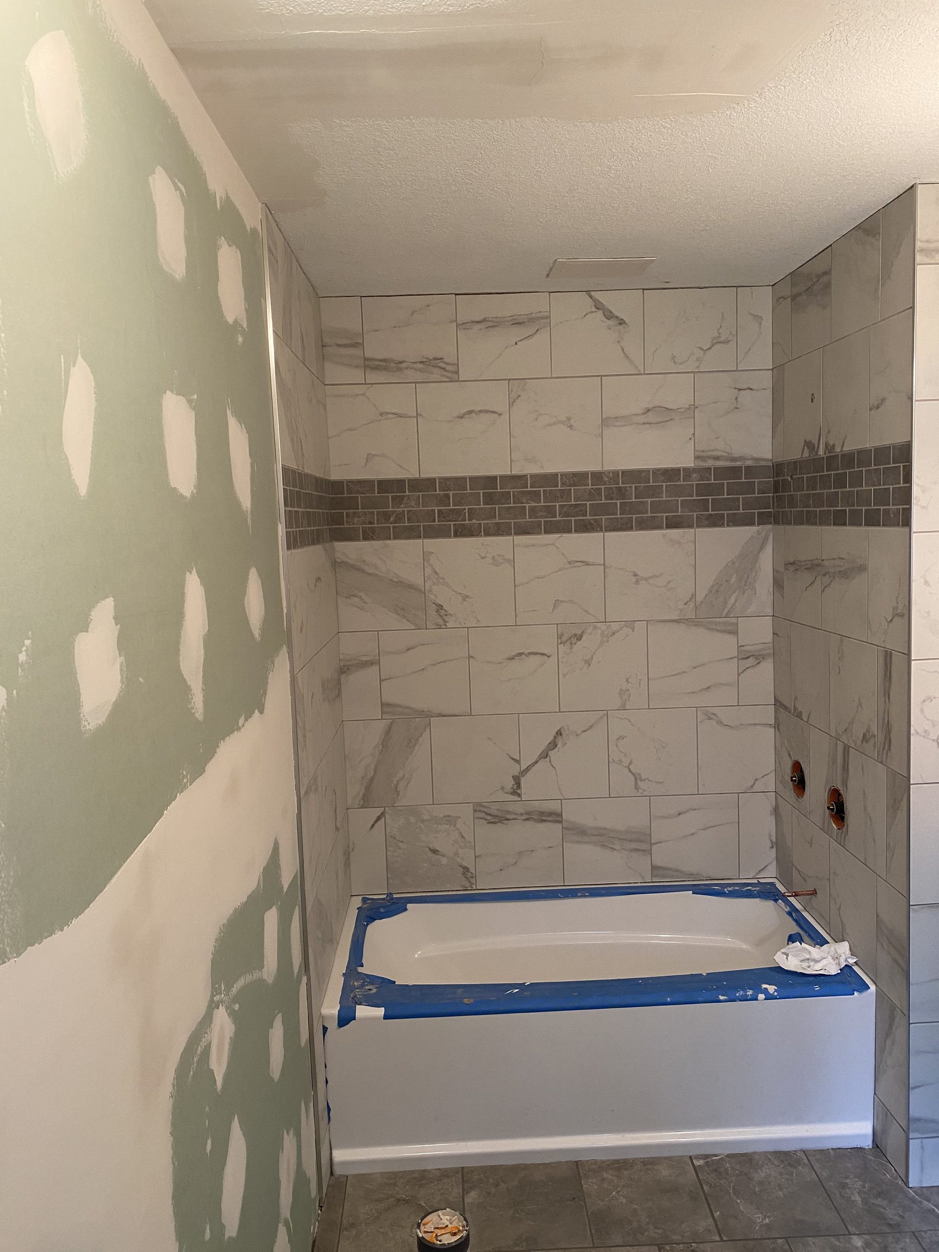 Tile shower enclosure installation in progress, showing marble-look ceramic tiles with dark accent band and white bathtub surround.